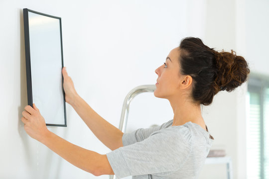young woman putting frame on white wall