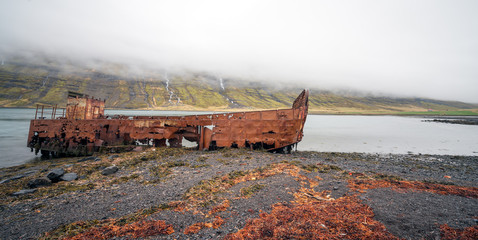 Rusty and abandoned old ship wreck from world war two laying on land in Mjoifjordur in Iceland. Waterfalls and foggy landscape scenery. Transport and ship concept.