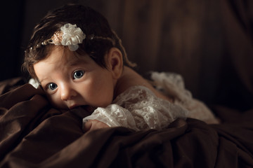 Beautiful infant in white dress, with flower headband lies on brown drapery, vintage blanket on wood background. Two month old girl. Surprised baby. Copy space