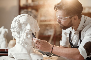 Professional bearded carver polishing with micro drill machine details of the man's sculpture head...