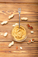 Peanut butter in spoon near creamy peanut paste in open glass jar, slice of peanut butter bread, toast. Peanuts in the peel scattered on the brown wooden table with copy space for cooking breakfast