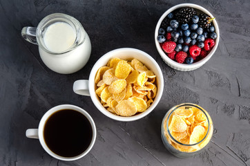 Healthy breakfast with cornflakes in a white cup, berries, milk and coffee on a dark gray background.