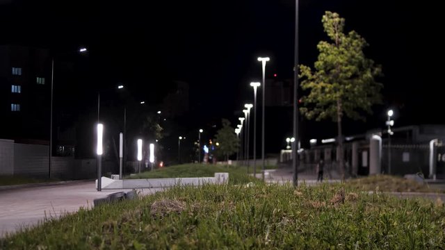 Public park infrastructure and night lighting. Stock footage. Man walking through green alley in the city park near bright street lamps.