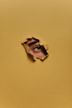 Eye of unrecognizable female model looking through ripped hole in yellow paper