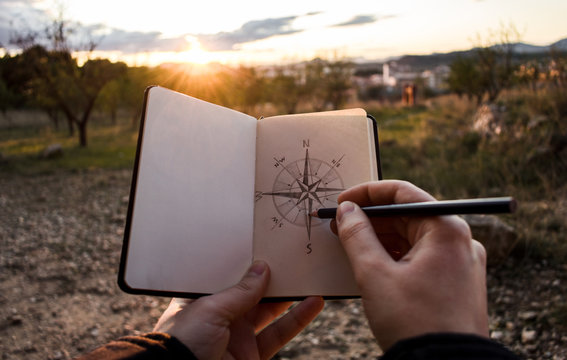Unrecognizable person drawing sketch of compass in notebook during trip through countryside in evening