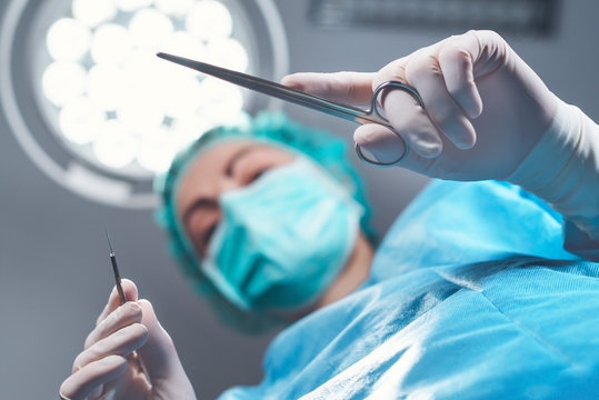 From below woman surgeon in medical uniform using professional tools while standing under bright light in operating theater