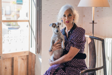 Pensive gray haired lady in elegant dress with obedient small Jack Russell Terrier dog sitting in vintage wooden armchair and looking at camera against light rustic interior