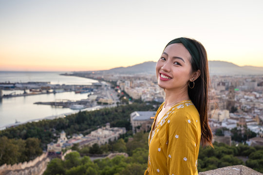 Side view of optimistic Asian lady smiling and looking at camera while visiting Castillo Gibralfaro on blurred background of coastal city and sundown sky in Malaga, Spain