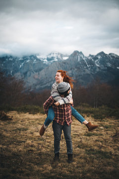 Lovely couple hugging and spending time together piggybacking and traveling in lawn with dry grass near by mountains in cloudy day