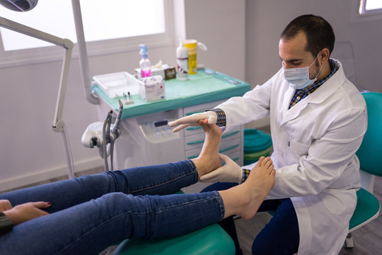 Side view of male podiatry doctor in white coat and protective mask massaging feet of female patient in modern medical center