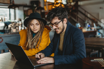 Smart man in glasses and stylish woman in hat looking at monitor while man typing on laptop in cafe