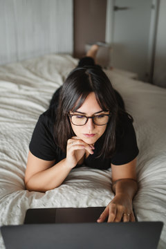 from above view of concentrated young woman in glasses typing on laptop while studying and laying on bed at home in Paris
