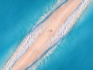 Aerial view of transparent blue sea with waves on the both sides and people on sandy beach at sunset. Summer holiday in Zanzibar, Africa. Tropical landscape with lagoon, white sand and ocean. Top view - obrazy, fototapety, plakaty