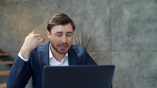 Smiling businessman chatting online on laptop camera in office. Guy vaving hand.