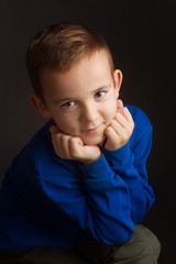 Studio portrait of a boy in a blue classic sweater on a dark background, propping up his chin with his fists