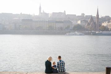 A young family of tourists sits on a stone parapet near the river and admires the beautiful Budapest. Sunny city.