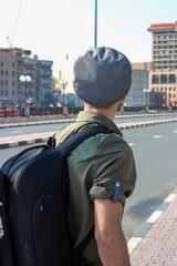 Back view of stylish young man in modern clothes with a hat and backpack close to the road