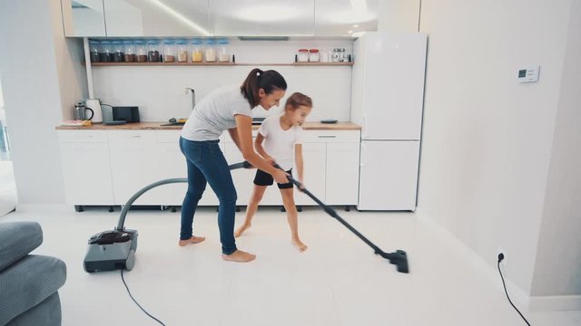 Woman with her daughter are vacuuming the kitchen floor with white tile without brush, only pipe of vacuum cleaner. Copy space. 4K.