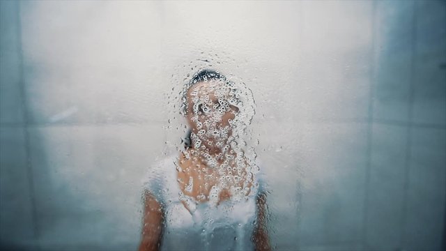 The woman washes a window in a bathroom. Splashes soapy water on the glass. Cleans the glass from dirt. Close up. 4K.