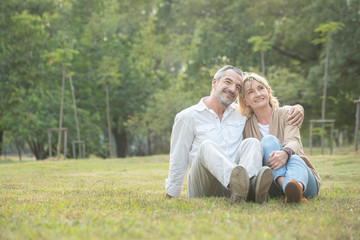 Fototapeta premium Senior elder caucasian couple sitting on ground together in park in Autumn. Wife resting head on husband head and put hands on man knee. Beautiful love relation and care of retirement old people.