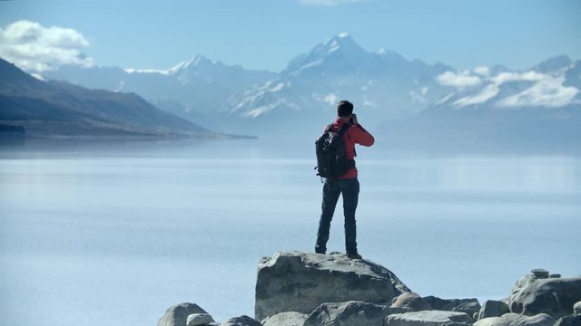 Mountain view, Lake Pukaki, New Zealand. Profile shot of young adventure photographer climbs atop a rock to take photos of the spectacular view with Mt Cook in distance. 10bit Pro Res