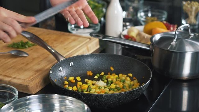 Close-up of cooking vegetables in kitchen, cook in pan brews corn, peas, onions and carrots, salt, pepper side dish, adds olive oil, steam from pan, stirs, table in kitchen is covered with pots