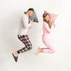 Jumping young couple in pajamas and with pillows on white brick background