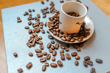 white mug full of coffee beans is standing on a white plate and grains are scattered around