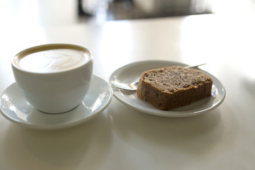 Fototapeta na wymiar Cappuccino in a white mug with a piece of cake on a white table in a cafe close-up.