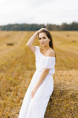 Fototapeta na wymiar A beautiful girl with blond hair in a white dress sits by a stack of straw in the field.