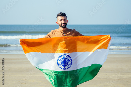 Indian young man waving indian tricolour flag outdoor near beach, conceptual image for republic day or independence day greeting
