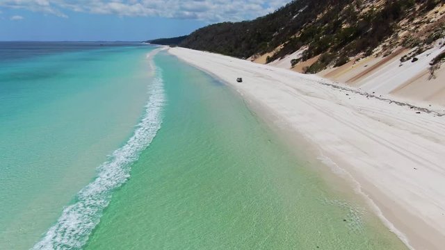 Stunning aerial drone footage of a white coloured four-wheel drive off-road vehicle driving along the vast beach on the West Coast of Fraser Island, Queensland, Australia.Clear water and gentle waves.
