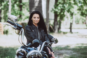 Obraz na płótnie Canvas Biker woman in a leather jacket on a motorcycle looks at the camera on a summer sunny day on a green background. Soft focus.