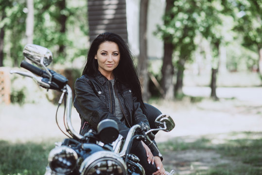 Biker woman in a leather jacket on a motorcycle looks at the camera on a summer sunny day on a green background. Soft focus.