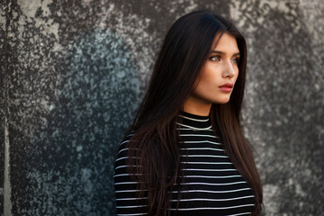 Fototapeta premium Beauty person. Young girl with straight brown hair, dressed in modern clothes, makeup, supported by a black wall, looking in profile.