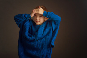 Studio portrait of dark-haired boy with a short haircut in a blue t-shirt and a knitted sweater, draped over his shoulders, holding his hands on his forehead, smiling. On a brown background