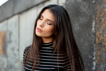 Beauty portrait of a sensual girl with straight brunette hair, dressed in modern clothes, with makeup, closed eyes, supported by a black wall.