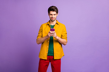 Fototapeta premium Photo of thoughtful concentrated man browsing through telephone in search of new information about red pants he bought isolated pastel violet color background