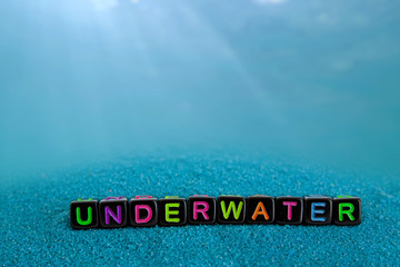 The word underwater is made up of colored letters on black cubes on blue sand