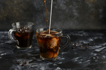   Two glass cups with cold coffee. Black iced coffee in transparent mugs. Streams of coffee and milk pour into a cup and intermix inside. Copy space