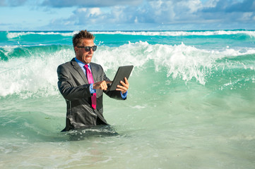 Shocked businessman trying to use his tablet computer in crashing waves on the shore of a tropical...