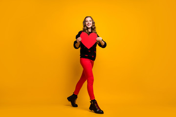 Fototapeta na wymiar Full length photo of pretty lady hold big paper heart walk street creative idea date invitation for boyfriend wear hearts pattern sweater red pants shoes isolated yellow color background