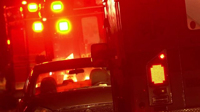 Car on side of the road between firetruck and ambulance with lights flashing at night.