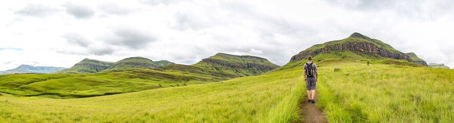 Man hiking on a small trail with a beautiful panorama of the green mountains of Maloti Drakensberg...