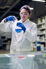 Young lab woman in glasses and white coat with flask with blue liquid in her hands conducts experiments