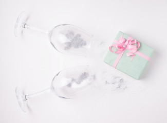 Gift in mint packaging on a white background next to glasses filled with sparkles. Holiday concept in light colors.