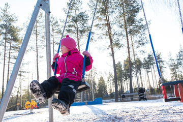 A child in winter clothes plays in the playground. The girl is riding on a swing. Winter entertainments in Finland.