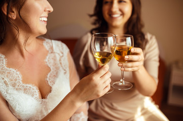 Beautiful smiling bride and bridesmaid drink wine at wedding morning.  Happy bride in white wedding dress with bridal bouquet at wedding morning. Newlywed woman with wedding makeup and hairstyle.