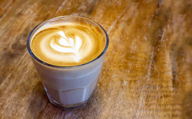 Hot cappuccino in glass on old wooden background. Coffee is decorated with flower pattern. Copy space.