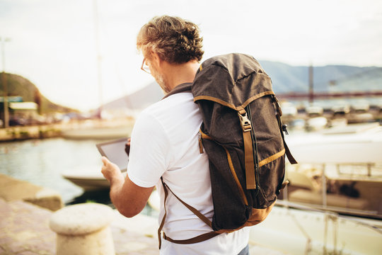Smiling tourist mature man standing with digital tablet and backpack near the sea.